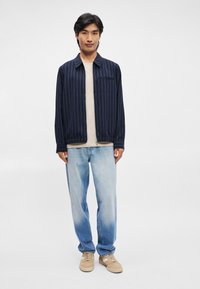 Ναυτική ριγέ ζακέτα με γιακά πάνω από μπεζ πουκάμισο, συνδυασμένη με ανοιχτό μπλε τζιν και καφέ αθλητικά παπούτσια. Η εμφάνιση είναι χαλαρή και άνετη.