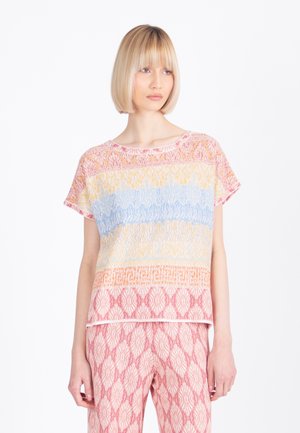 Femme aux cheveux blonds coiffés en carré, portant un haut à manches courtes avec des motifs horizontaux pastel et un pantalon rose à motifs, debout devant un fond blanc.