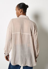 Blusa de gasa color beige con rayas verticales, cuello y mangas largas, que presenta un corte holgado y una tela ligeramente texturada.