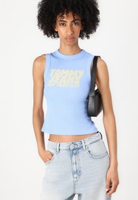 Top corto a coste azzurro chiaro con il testo in giallo audace "TOMMY JEANS NEW YORK CITY". Abbinato a pantaloncini di jeans a vita alta e una piccola borsa nera.