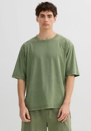 Joven con cabello corto y oscuro que lleva una camiseta verde holgada y pantalones cortos a juego, posando frente a un fondo liso y claro.