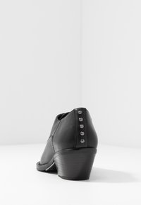 Bottine en cuir noir avec un talon empilé, ornée de clous métalliques le long de l'arrière et une texture lisse. Vue latérale sur fond blanc.