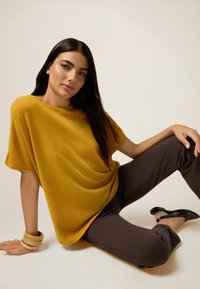 Jersey oversized de color mostaza, con textura suave, combinado con unos pantalones ajustados marrón oscuro. El modelo lleva una pulsera de bangle multicolor y zapatos negros.