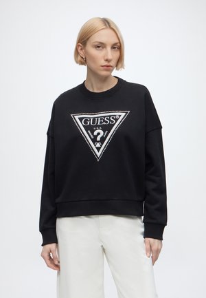 Donna bionda che indossa una felpa nera Guess con un grande logo triangolare e pantaloni bianchi, in piedi davanti a uno sfondo chiaro e uniforme.