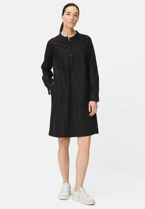 Vrouw staand met één hand in haar zak, gekleed in een zwarte, knoopjurk met lange mouwen tot op de knie en witte sneakers, tegen een witte achtergrond.