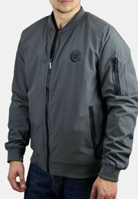 Chaqueta bomber gris con cierre de cremallera en el frente, puños y collar acanalados. Cuenta con bolsillos laterales con cremallera y un pequeño logotipo en el pecho. Textura suave.