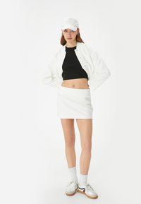 Witte bomberjack over een zwart crop top, gecombineerd met een witte minirok. Witte pet, witte sportsokken en witte sneakers met accenten.