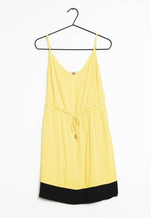 Vestido amarillo sin mangas con tirantes ajustables, cintura con cordón y dobladillo negro colgado en una percha negra contra un fondo blanco.