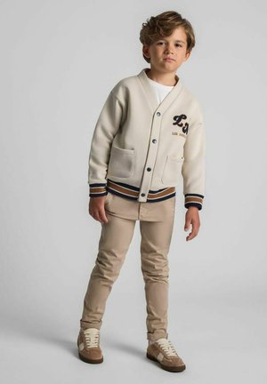 Junge mit hellbraunen Haaren trägt eine beige Collegejacke, beige Hosen und braune Sneakers und steht vor einem schlichten grauen Hintergrund.