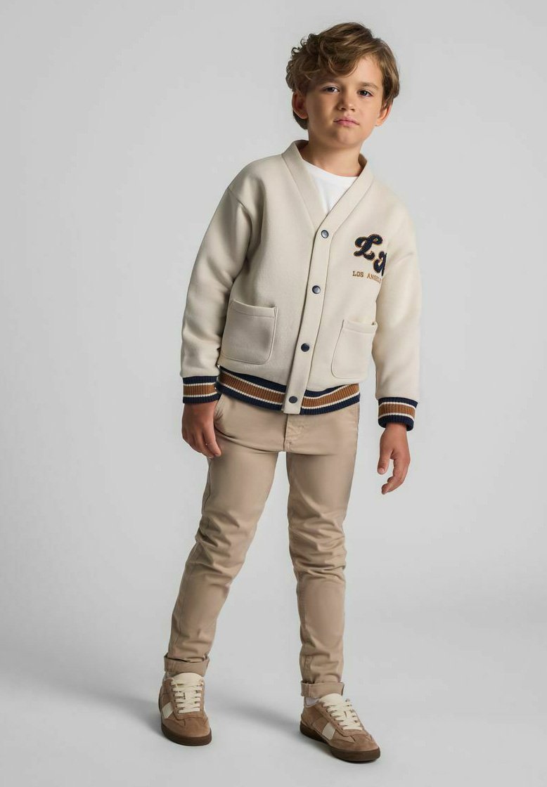 Berniukas su šviesiai rudais plaukais vilki smėlio spalvos sportinį švarką, smėlio spalvos kelnes ir rudus sportbačius, stovintis prieš pilką vientisą foną.