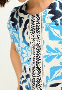 Blusa con estampado de grandes diseños florales azules sobre un fondo blanco, con un acento vertical negro en el centro que contrasta.