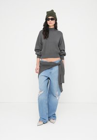 Πλεκτό πουλόβερ σε γκρι χρώμα με μεγάλο μέγεθος, ανοιχτό μπλε τζιν παντελόνι με φαρδιά μπατζάκια και φθορές, γκρι δέσιμο στη μέση, πράσινο σκούφο και μαύρα γυαλιά ηλίου.