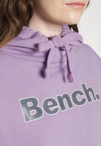Zbliżenie na osobę w fioletowej bluzie z kapturem z związanymi sznurkami i nadrukiem "Bench." dużymi czarnymi i białymi literami na klatce piersiowej.