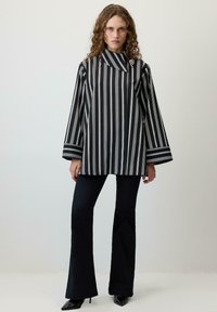 Ženska s kodrastimi lasmi nosi črno-belo zgornji del z navpičnimi črtami, črne razširjene hlače in črne ostro konične pete, stoji ob običajni steni.