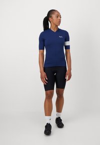 Marine sykkelskjorte med korte ermer og glidelås foran, med en hvit aksentstripe. Kombinert med svarte sykkelshorts og sko.