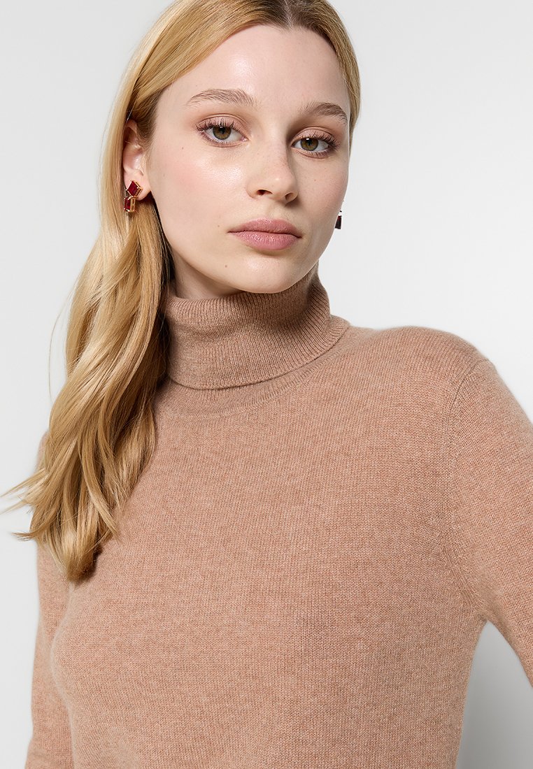 Beżowy sweter z golfem wykonany z miękkiej, teksturowanej tkaniny. Dopasowany krój z prążkowanym kołnierzem. Modelka nosi kwadratowe kolczyki.
