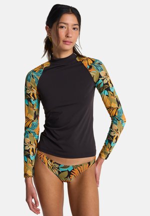 Jeune femme portant un rash guard noir avec des manches florales tropicales multicolores et un bas de bikini assorti, debout devant un fond uni.