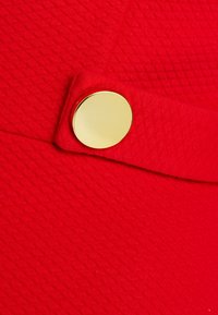 Punainen kangas, jossa on tekstuurinen kuvio ja kultainen pyöreä nappi yksityiskohtana. Nappi on kiinnitetty vaakasuoraan nauhaan.