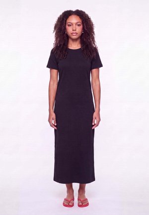 Femme aux cheveux bouclés portant une robe longue noire à manches courtes et des tongs rouges, debout devant un fond clair uni.