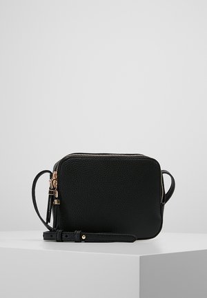 Sac bandoulière rectangulaire en cuir noir texturé avec fermeture éclair dorée et bandoulière ajustable sur surface blanche contre un fond gris.
