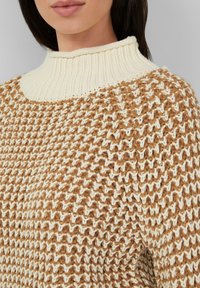 Frau in einem grob gestrickten Pullover mit hohem, geripptem cremefarbenem Kragen und einem braun-cremefarbenen strukturierten Muster.