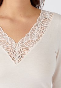 Top beige con scollatura a V profonda, caratterizzato da intricate rifiniture in pizzo bianco, che presenta un motivo testurizzato lungo il collo. Tessuto morbido.