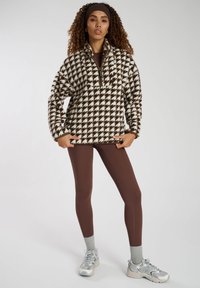 Pullover in pile a scacchi marrone e bianco con zip a metà, abbinato a leggins marroni e calzini grigi, completato da sneakers grigie.