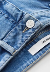 Gros plan d'un jean en denim bleu montrant un bouton métal avec un logo et une étiquette de marque en tissu blanc partiellement visible cousue à l'intérieur de la ceinture.