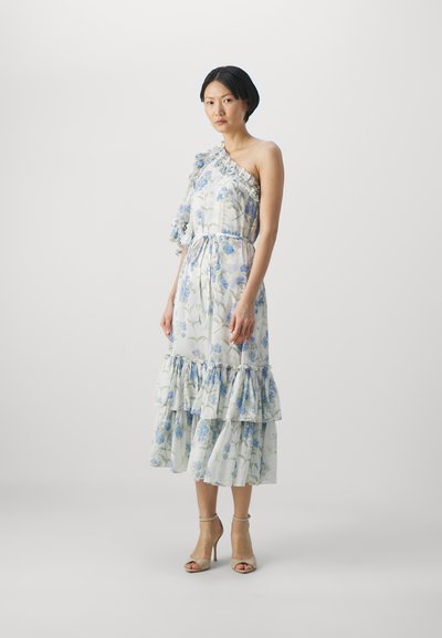 Witte bloemenjurk met blauwe bloemen, ruches op één schouder, volantzoom en een ceintuur in de taille. Voltooid met beige hoge hakken.