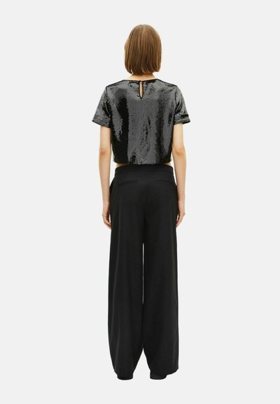 Donna che indossa un crop top nero a maniche corte con paillettes e pantaloni neri a vita alta e gamba larga, in piedi di spalle contro uno sfondo bianco.