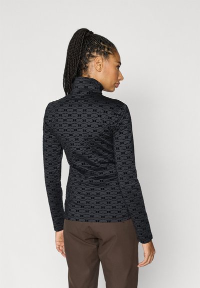 Melns augsts kakls ar teksturētu atkārtotu rakstu. Garas piedurknes un pieguļošs siluets, kombinēts ar brūnām biksēm.