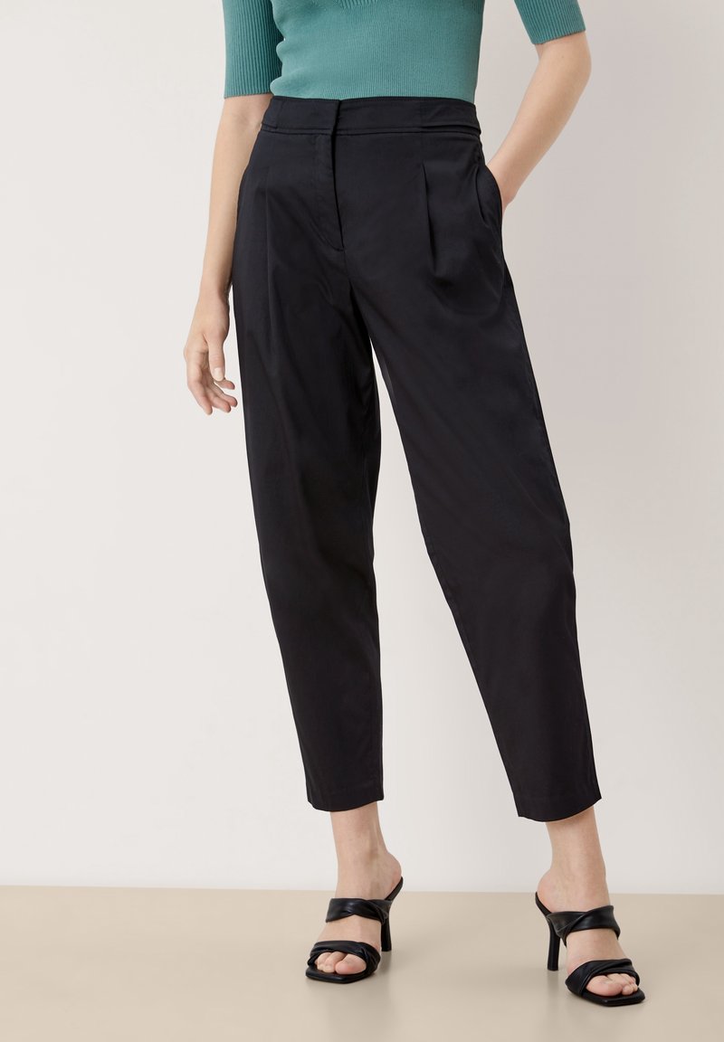 Pantalon noir fuselé avec des plis, fabriqué en tissu lisse. Porté avec des mules à talons ouvertes noires. Présenté sur un fond neutre.