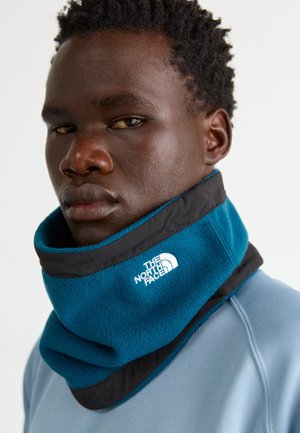 Mann trägt eine blaue und schwarze Fleece-Nackenwärmer mit dem The North Face-Logo, gekleidet in ein hellblaues Oberteil vor einem einfarbigen Hintergrund.