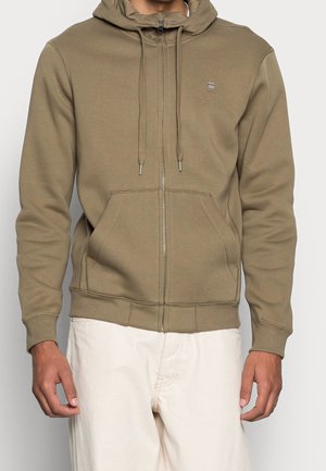 Homme portant un sweat à capuche zippé marron clair avec poches avant et cordons, associé à un pantalon blanc cassé sur fond uni.