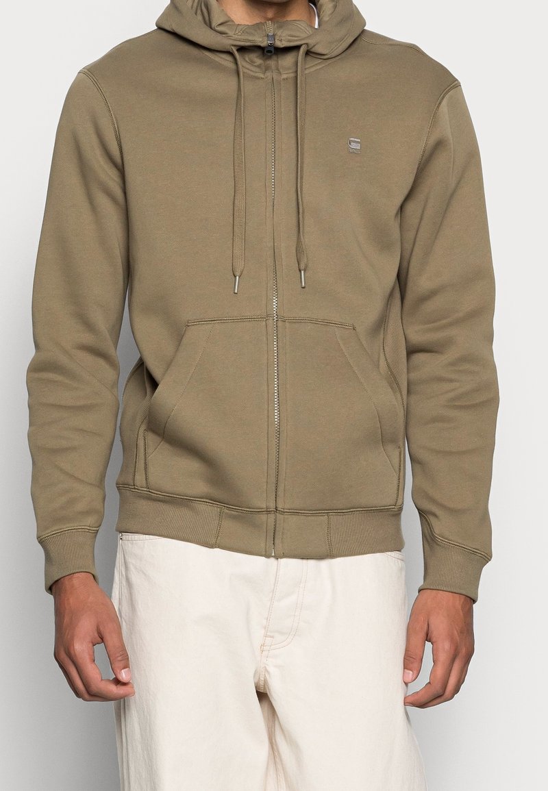 Homme portant un sweat à capuche zippé marron clair avec poches avant et cordons, associé à un pantalon blanc cassé sur fond uni.