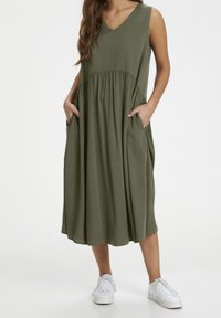 Robe sans manches vert olive avec encolure en V, taille froncée et poches latérales ; tissu doux avec une coupe ample, associée à des baskets blanches.