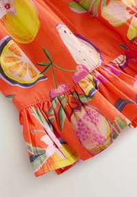 Klart orange tyg med färgglada frukttryck inklusive körsbär, citronskivor, ananas och blommor, med en rynkad volangkant.