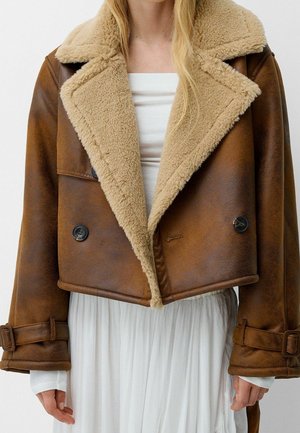 Frau trägt eine kurze braune Lederjacke mit beigem Lammfellkragen und Manschetten über einem weißen Oberteil und einem fließenden Rock.