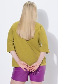 Top verde oliva de manga 3/4 combinado con pantalones cortos morados brillantes; el cabello es lacio y largo, con las manos colocadas detrás de la espalda. Fondo minimalista.