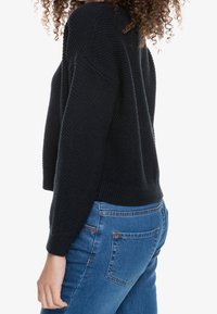 Suéter negro de punto con un patrón texturizado, diseño corto y puños de canalé, combinado con jeans azules de tiro medio que cuentan con bolsillos traseros.