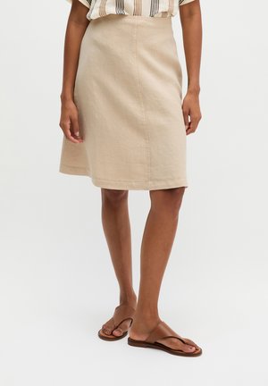 Vrouw die een beige knielange rok draagt, een gestreept topje met korte mouwen en bruine platte sandalen, staand tegen een effen witte achtergrond.