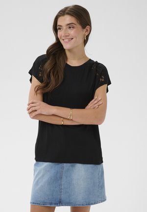 Junge Frau mit langen braunen Haaren, trägt ein schwarzes Oberteil mit Spitzensäumen, goldenen Schmuck und einen hellblauen Jeansrock, Arme verschränkt, lächelt.