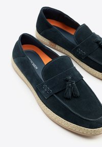 Marineblaue Suede-Live mit Quasten-Details, kontrastierendem orangenem Innenraum und einer geflochtenen Jutesohle. Glatte Textur und klassisches Design.