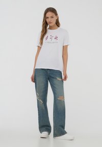 T-shirt bianco in cotone con stampa floreale e testo "LE JARDIN PARISIEN", abbinato a jeans larghi distressed blu chiaro e sneakers bianche.