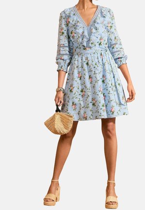 Femme portant une robe fleurie bleu clair avec des détails à volants, tenant un sac à main tressé et portant des sandales à plateforme beige.