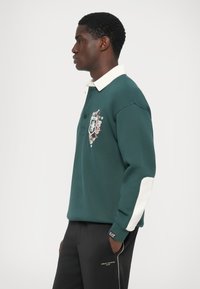 Zelena sweatshirt z belim ovratnikom, teksturirana tkanina, cvetlični detajli in aplikacija. Črne hlače z logotipom in belimi poudarki.