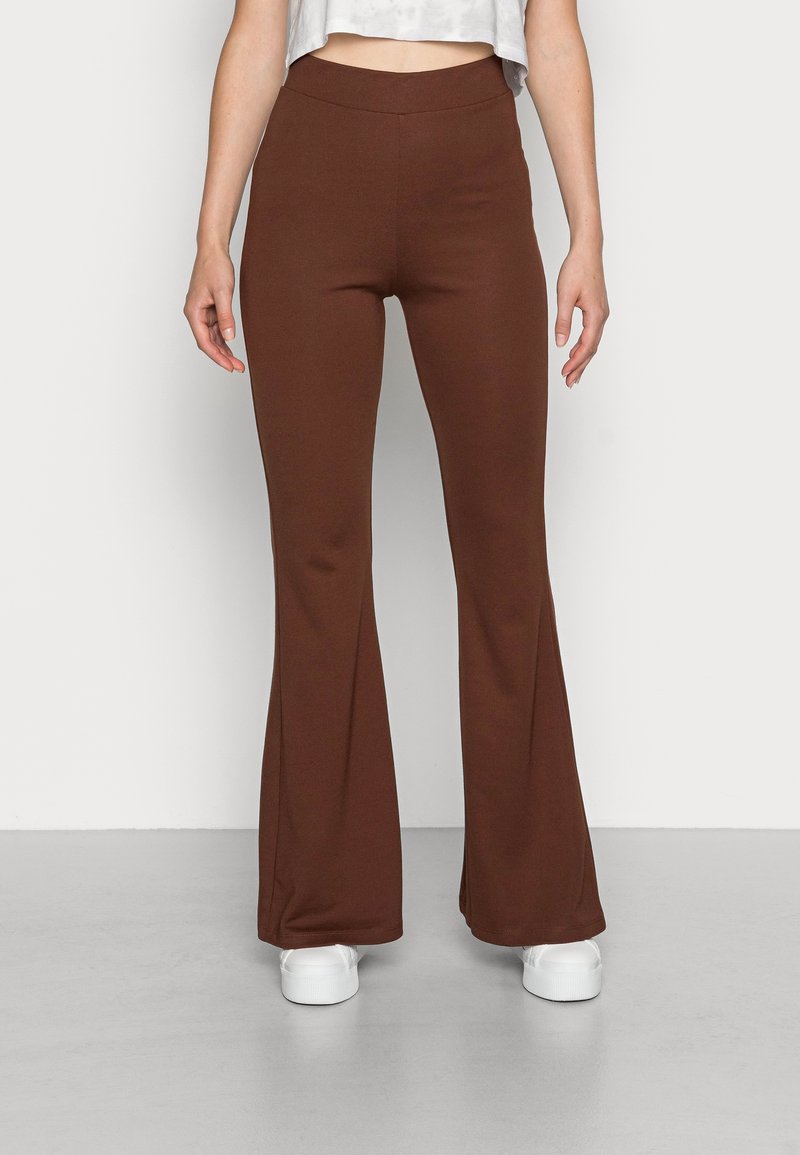 Femme portant un pantalon marron évasé et des baskets blanches, debout contre un mur blanc uni, mains détendues le long du corps.
