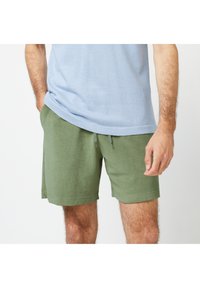 Shorts verts en mélange de coton texturé, dotés d'une taille élastique avec cordon de serrage et de poches latérales. Portés avec un t-shirt bleu clair.