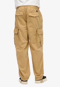 Un pantalon cargo beige présente une taille élastique, plusieurs poches et des poignets ajustables, avec une texture douce et un design décontracté.