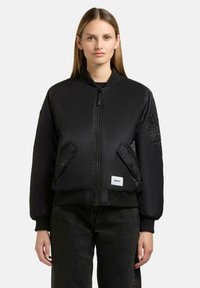 Schwarze Bomberjacke mit Reißverschluss vorne, gerippten Bündchen und zwei schrägen Taschen. Mit einem Logo-Patch auf der unteren linken Seite und einer Taschen an den Ärmeln.